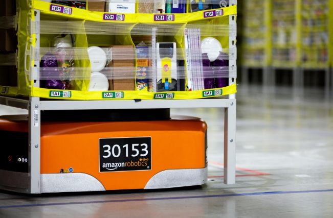 Photograph of orange Amazon Robotics drive units carrying storage shelves to a picking station.